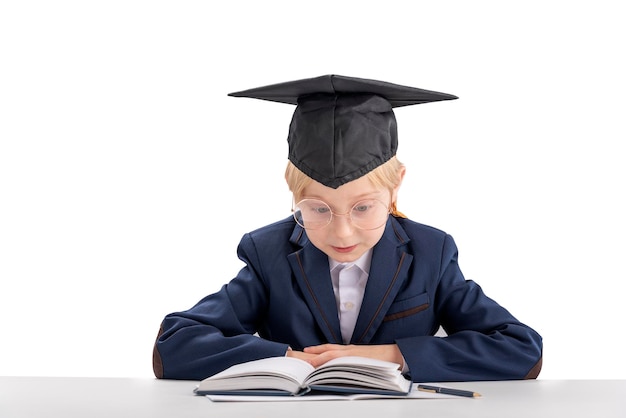 Niño con gafas grandes viste uniforme escolar y sombrero de estudiante sentado en la mesa leyendo libros de texto aislado sobre fondo blanco.