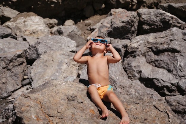 Un niño con gafas de sol está sentado y tomando el sol en grandes rocas en la playa cerca del mar