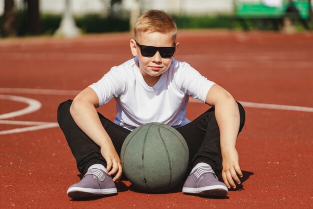 Niño con gafas de sol se sienta en una cancha de baloncesto con una pelota en verano. Foto de alta calidad