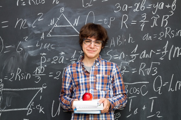 Niño con gafas sosteniendo libros con manzana, pizarra llena de fórmulas matemáticas antecedentes