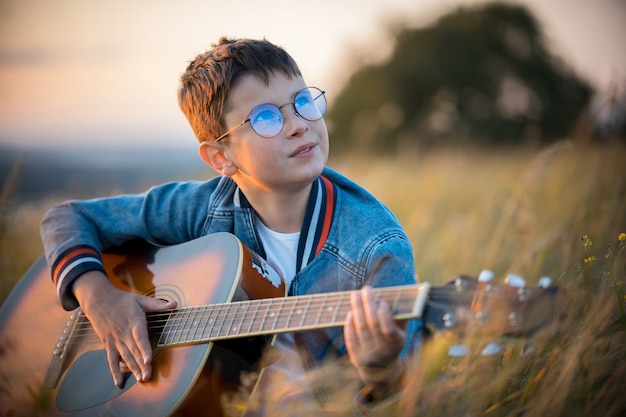 Un niño con gafas tocando la guitarra en el campo. Belleza natural.