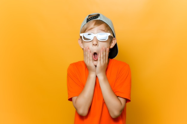 Un niño con gafas para ver películas abrió la boca con sorpresa y se sujeta las mejillas con las manos.