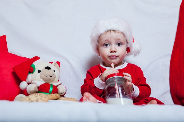 Un niño con una gorra roja come galletas y leche. Fotografía navideña de un bebé con una gorra roja.