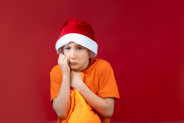 Un niño con un gorro de Papá Noel de Año Nuevo sostiene su mano cerca de su rostro y presiona una bolsa de regalo para sí mismo