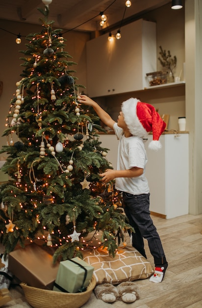 Foto un niño con un gorro de papá noel decora un gran árbol de navidad en casa. concepto de navidad.