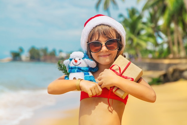 Foto niño con gorro de papá noel y gafas de sol con regalos en la playa.