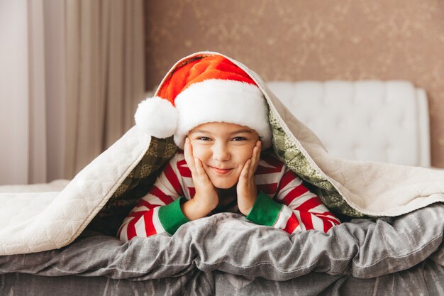 Niño con un gorro de Papá Noel y pijama se encuentra en una cama con sábanas blancas, año nuevo, navidad