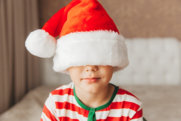 Niño con un gorro de Papá Noel y pijama se encuentra en una cama con sábanas blancas, año nuevo, navidad