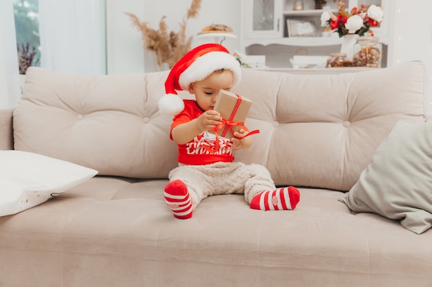 Un niño con un gorro de Papá Noel se sienta en el suelo cerca del sofá y sostiene una caja con un regalo en sus manos. Navidad