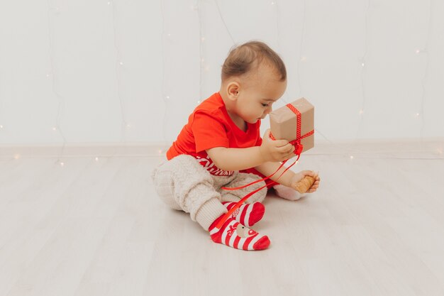 Un niño con un gorro de Papá Noel se sienta en el suelo cerca del sofá y sostiene una caja con un regalo en sus manos. Navidad