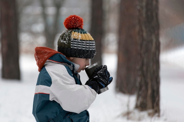 Niño con gorro de punto descansando y bebiendo té caliente o cacao del termo en el frío día de invierno en el bosque.