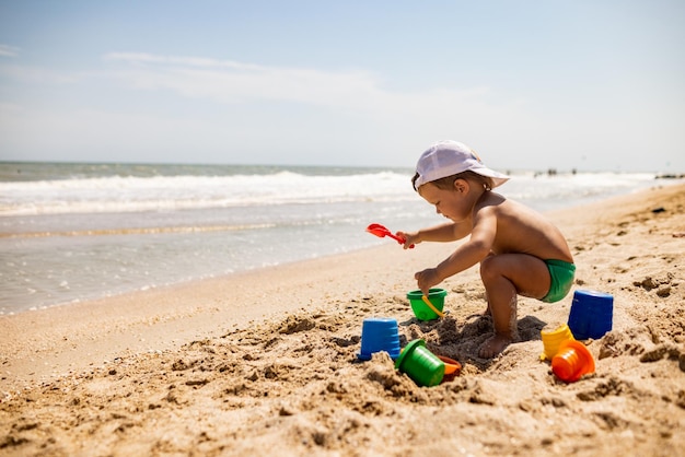 Un niño gracioso y divertido recoge conchas y guijarros en el mar azul tranquilo en un fondo arenoso bajo el sol de verano en unas vacaciones brillantes