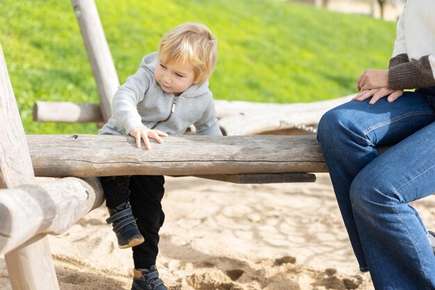 Un niño gracioso jugando en el patio de recreo con troncos de madera bajo la supervisión de su madre