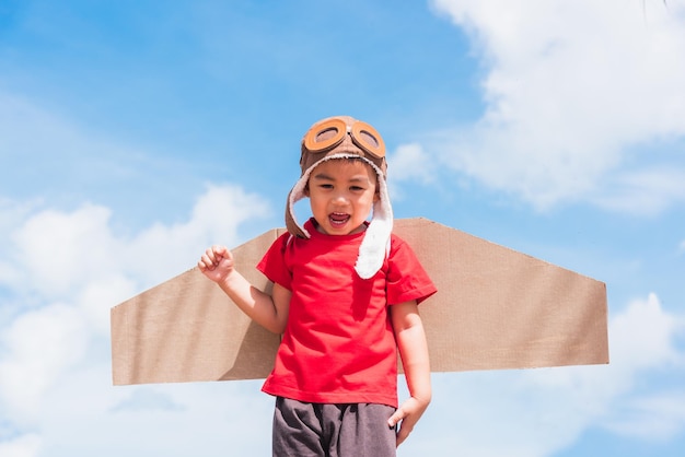 Niño gracioso, niño pequeño, sonrisa, lleva sombrero de piloto y gafas de protección, juega a las alas de un avión de cartón de juguete.