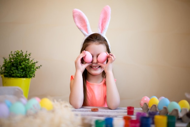 Niño gracioso sostiene huevos de pascua cerca de los ojos preparándose para pascua con orejas de conejo