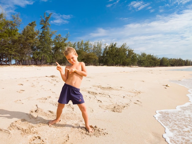 Un niño guapo baila en la playa sobre arena limpia bajo un cielo azul y disfruta de las vacaciones en el mar Infancia feliz y concepto de turismo con niños