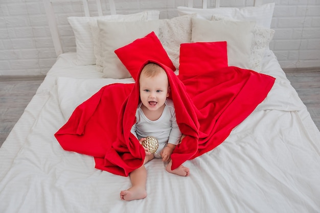 niño guapo con un traje blanco está sentado en una cama con una manta roja