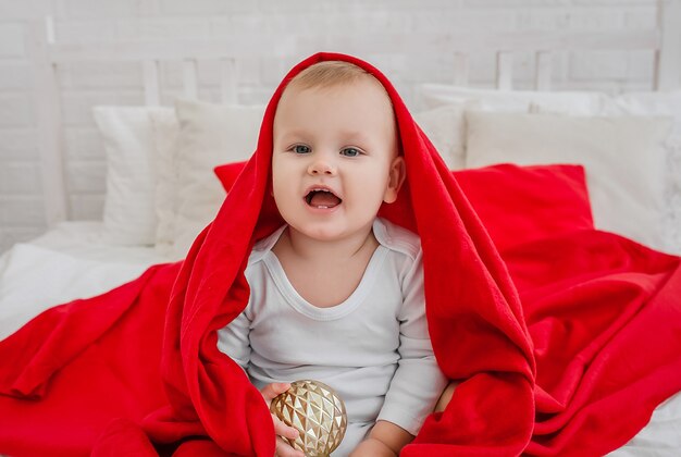 niño guapo con un traje blanco está sentado en una cama con una manta roja