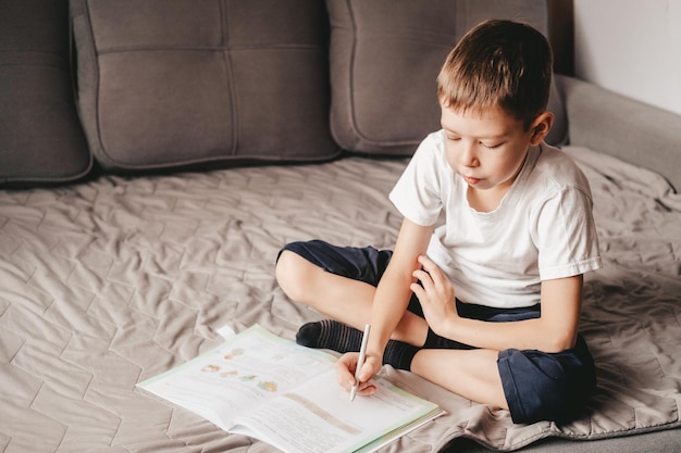 El niño hace la tarea mientras está sentado en el sofá gris. Un adolescente caucásico escribe en un libro en el sofá. Educación en el hogar de un niño de 9 años. Aprendizaje libre