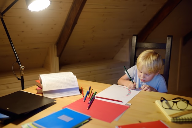 Niño haciendo los deberes, pintando y escribiendo en la noche de hogar. Los niños en edad preescolar aprenden lecciones: dibujar y colorear imágenes. Entrenamiento para niños para escribir y leer.