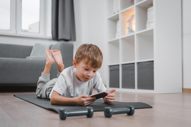 Niño haciendo ejercicio con pesas en casa, eligiendo una lección en el teléfono inteligente, viendo videos de fitness en Internet o teniendo una clase de fitness en línea, interior de la sala de estar.