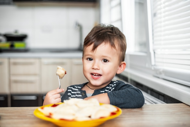 Niño hambriento comiendo bolas de masa en la cocina, sentado a la mesa con una chaqueta gris