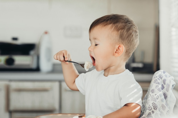 Un niño hambriento está comiendo bolas de masa en la cocina sentado en una silla con una camiseta blanca