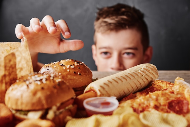 Niño hambriento mira comida sabrosa y toma una hamburguesa de la mesa