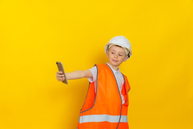 Un niño hermoso vestido con un casco y un chaleco reflectante hace un selfie en un teléfono inteligente, se encuentra en una pared amarilla.