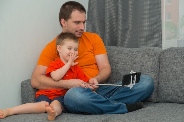 Niño y hombre en ropa brillante mirando en el teléfono inteligente durante la llamada en línea y sonriendo. Concepto de comunicación a distancia y en casa