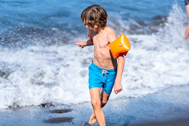 Foto niño huyendo de las olas del mar. el niño lleva mangas de brazo y se divierte en el agua.