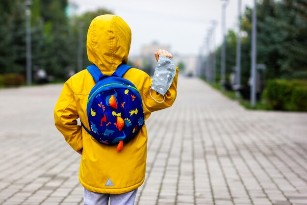 Foto un niño con un impermeable amarillo con una mochila sosteniendo una máscara médica, la escuela está en autoaislamiento