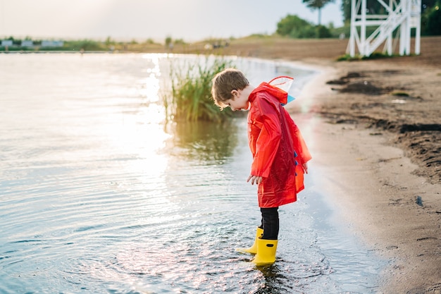 Niño con un impermeable rojo y botas de goma amarillas jugando con agua en la playa, niño de la escuela en un