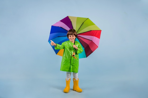 Un niño con un impermeable verde y botas de goma amarillas se encuentra con un paraguas multicolor se encuentra sobre un fondo azul con un lugar para el texto