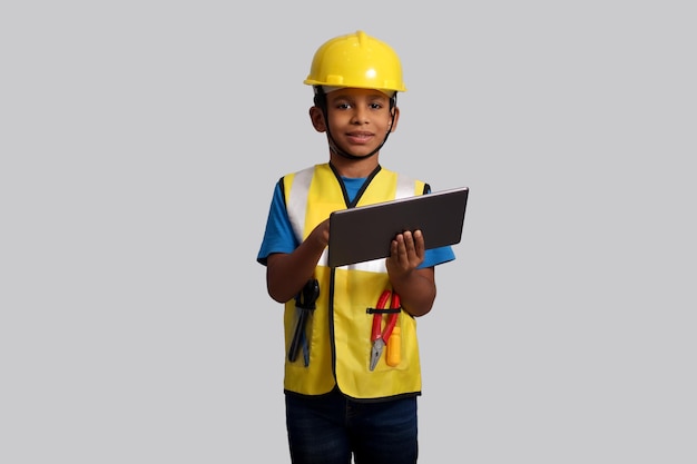 Niño indio de 7 a 8 años con casco amarillo y chaqueta de seguridad con una tableta en la mano