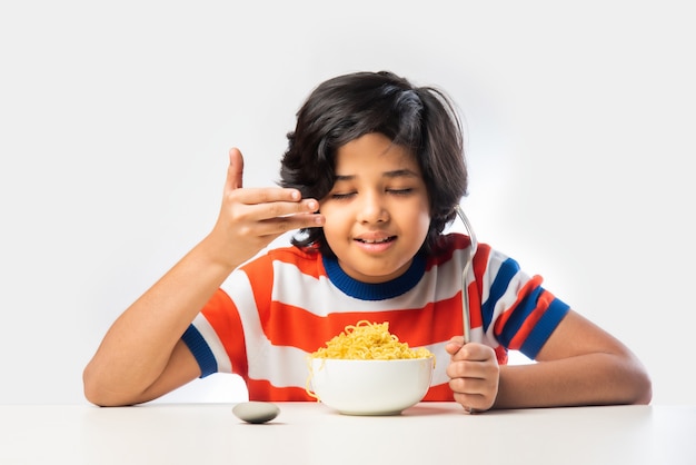 Niño indio comiendo deliciosos fideos con tenedor contra el fondo blanco, niño asiático come espaguetis en un recipiente contra el fondo blanco.