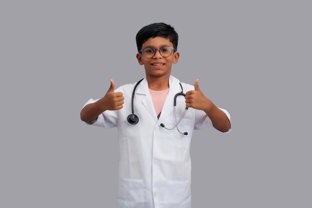 Foto niño indio con un delantal de médico con un estetoscopio de pie y mostrando los pulgares con las manos