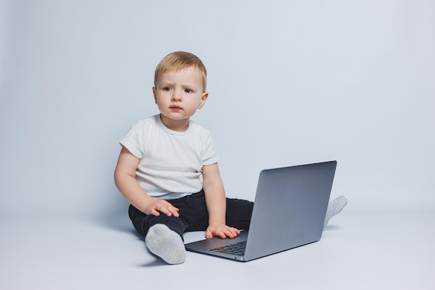 Un niño inteligente de 34 años se sienta con una computadora portátil sobre un fondo blanco Un niño con una camiseta blanca y pantalones negros se sienta en una computadora portátil y mira a la cámara Niños modernos