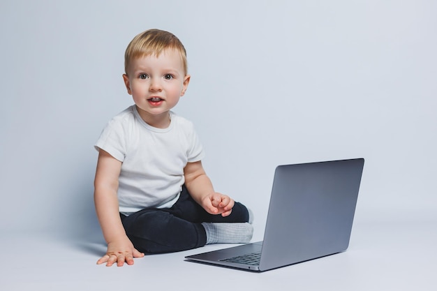 Un niño inteligente de 34 años se sienta con una computadora portátil sobre un fondo blanco Un niño con una camiseta blanca y pantalones negros se sienta en una computadora portátil y mira a la cámara Niños modernos