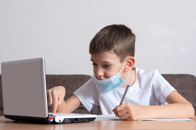 Un niño inteligente, un colegial, está sentado en un escritorio en casa, estudiando en línea en una computadora portátil y escribiendo información en su cuaderno. Concepto de educación en línea, aprendizaje a distancia