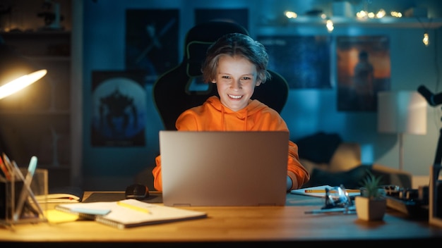 Niño inteligente usando una computadora portátil en una habitación oscura acogedora en casa Adolescente feliz navegando por la investigación educativa en línea Charlando con amigos Estudiando tareas escolares