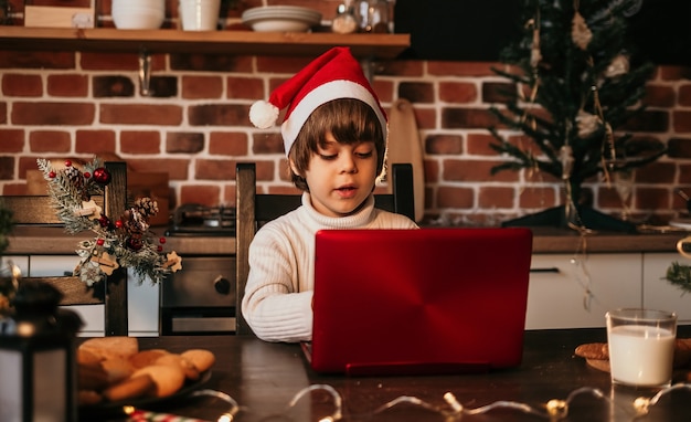 Un niño interesado en Año Nuevo con un suéter blanco y un sombrero rojo de Año Nuevo está sentado en una mesa y escribiendo una carta en una computadora portátil roja