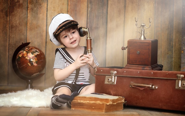 Un niño en un interior retro y un teléfono viejo se sienta en el suelo. Un niño pequeño, un viajero con decoraciones vintage. El niño viajero está llamando por teléfono.