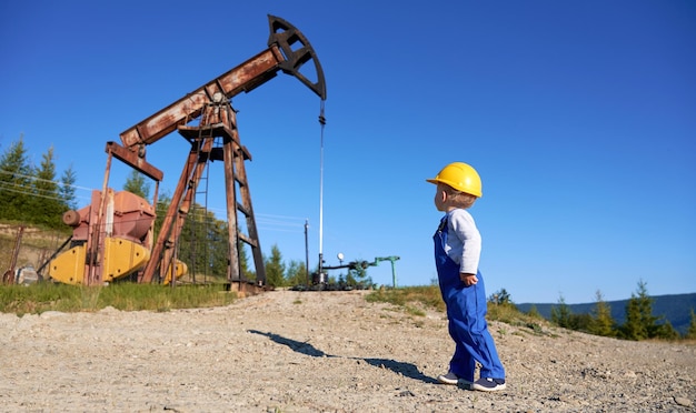 Foto niño joven con mono azul y casco amarillo mirando una máquina en alta mar en un entorno natural y pensando en la educación técnica sistema de aprendizaje masculino de plataforma de perforación y bomba de minería