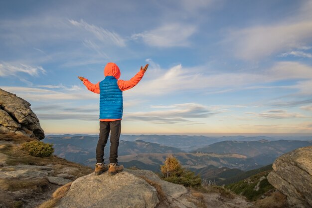 Niño joven muchacho excursionista de pie con las manos levantadas en las montañas disfrutando de la vista del increíble paisaje de montaña al atardecer.
