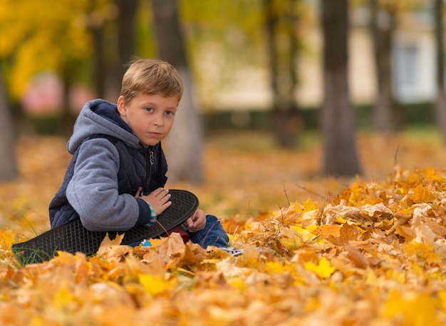 Un niño joven y solitario con su patineta sentado en hojas de otoño marrón en un parque.