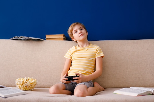 niño juega con gamepad en lugar de lecciones y come palomitas de maíz