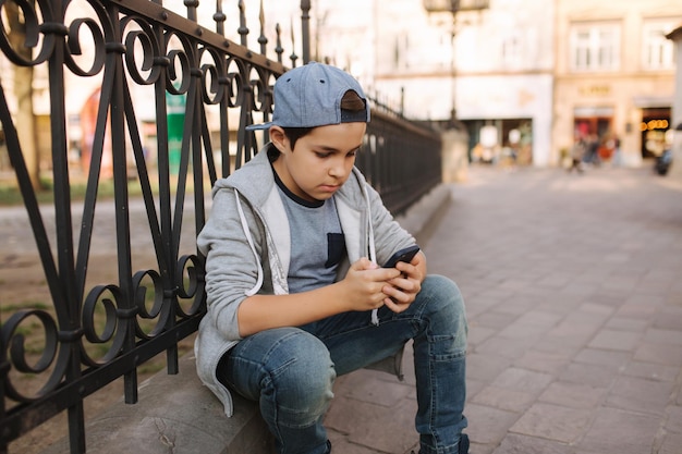 Niño juega en línea en el teléfono inteligente afuera Niño sentado en el centro de la ciudad Niño juega mientras su madre va a la tienda