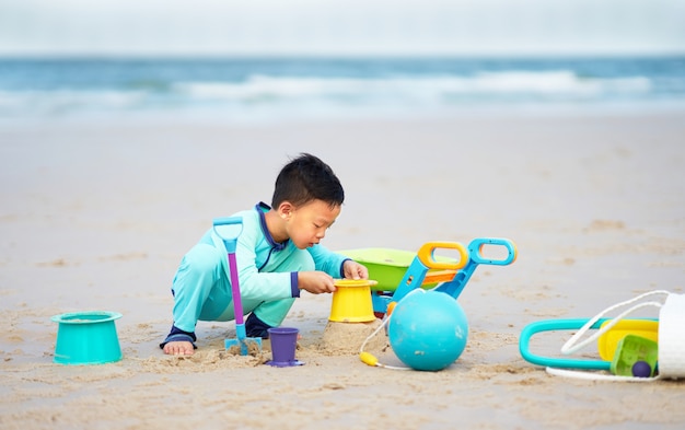 Niño juega pala y juguetes en la arena de la playa solo con feliz en concepto de vacaciones de verano con espacio de copia