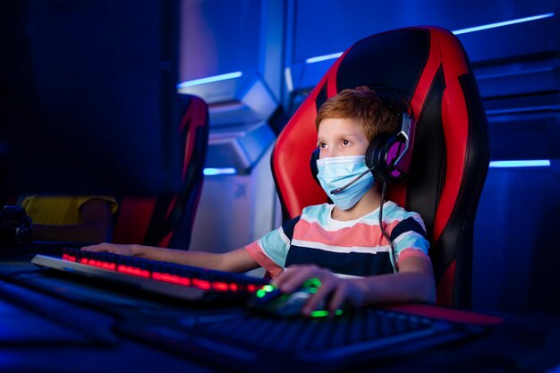 niño jugador con mascarilla y auriculares jugando videojuegos en la computadora en la sala de juegos